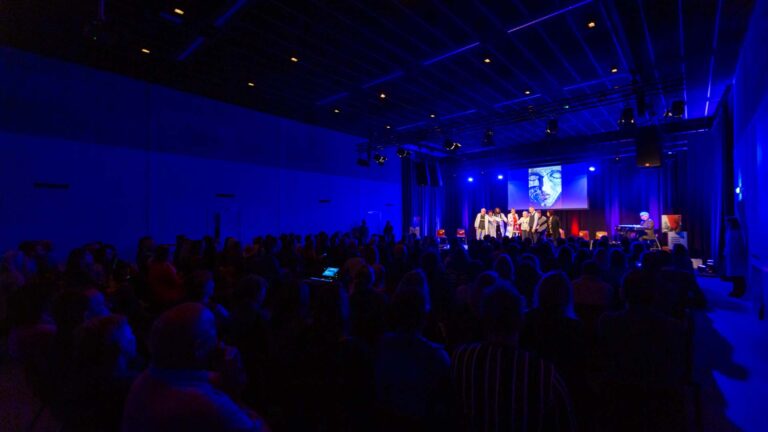 De programmazaal gefotografeerd tijdens een evenement met een aantal personen op het podium, muzikale begeleiding van een pianist en een presentatie op het scherm achter het podium